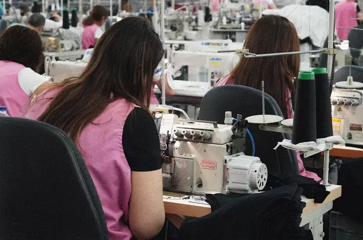 Costureras con batas rosas cosiendo prendas negras en máquinas remalladoras